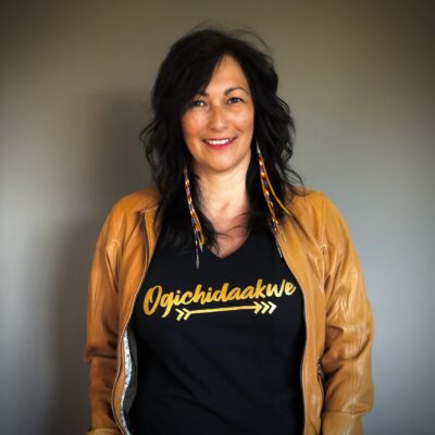 A photo of indigenous innovation expert Diane Roussin, a smiling woman with long, dark hair standing in front of a grey background wearing a yellow jacket 